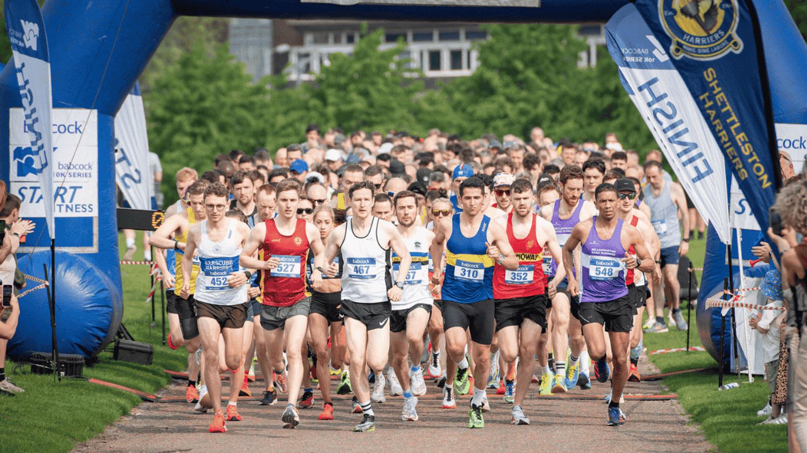 A large group of runners in colorful athletic gear start a race from beneath a blue inflatable arch on a paved path. The scene is set outdoors with trees and spectators in the background.