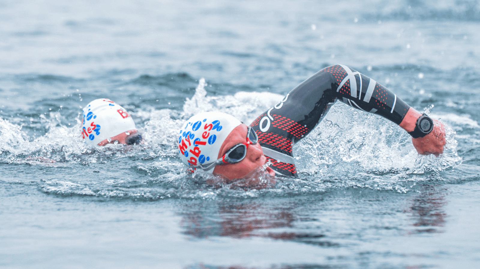Two swimmers wearing white caps with blue and red logos swim in open water. The foreground swimmer wears a wetsuit and goggles, with a watch on their wrist, performing a front crawl stroke. The background swimmer follows closely.