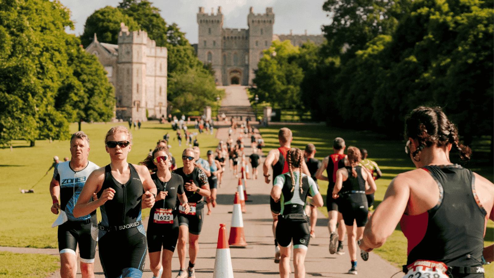 A group of athletes in triathlon gear running along a wide road lined with orange cones and lush trees. In the background, a large, historic castle with towers and an arched entrance is visible. Spectators watch from the grass on either side.
