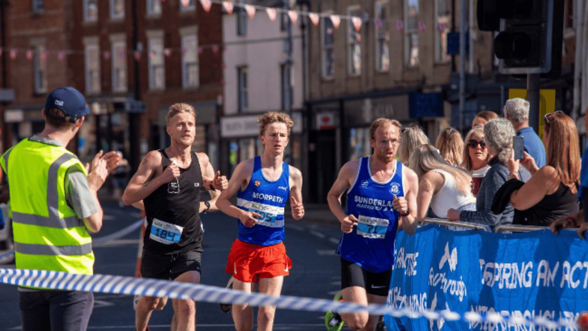 Three runners compete in a race, passing by spectators and a volunteer in a fluorescent vest. They are on a city street, with shops and buildings in the background. A banner and tape line the route.