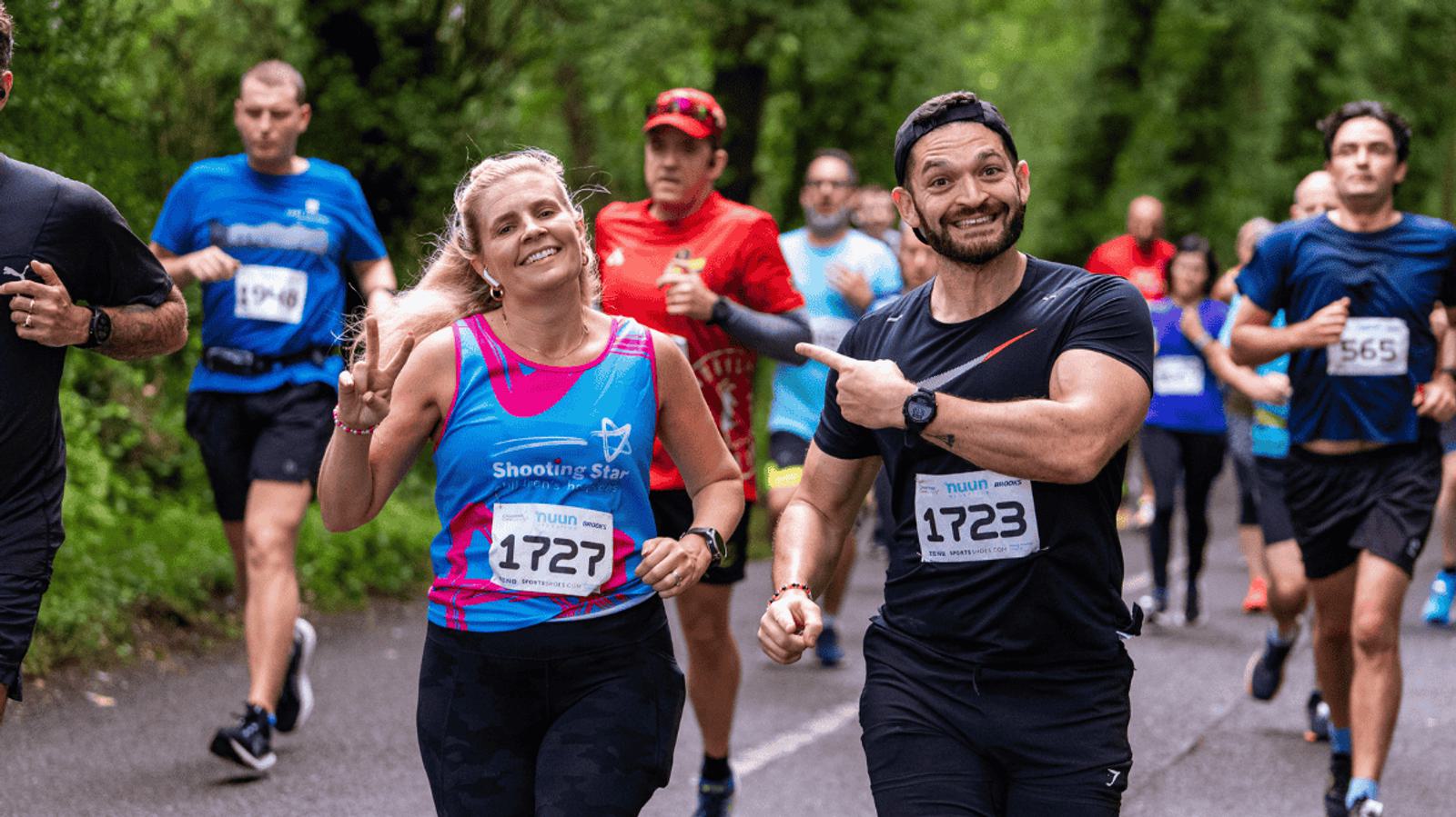 A group of runners participating in a race on a tree-lined road. A man in a black outfit points playfully at a smiling woman in a pink and blue tank top. They're both wearing race bibs, surrounded by other runners in the background.