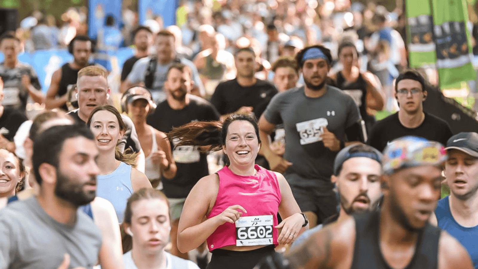 A large group of runners participating in a race on a sunny day. A smiling woman in a pink tank top and wearing number 6602 is in the foreground, with other runners surrounding her. Trees and sponsor banners are visible in the background.