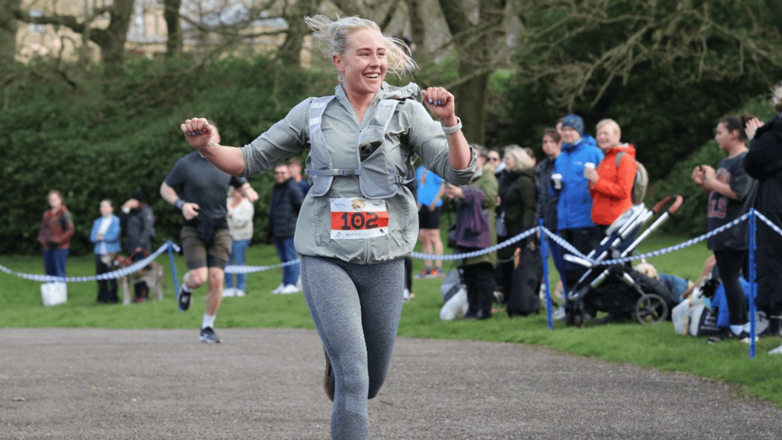 A woman in athletic gear smiles as she crosses a race finish line. She is wearing a gray vest and leggings. Spectators with strollers and folding chairs watch and cheer from the sidelines in a park setting.
