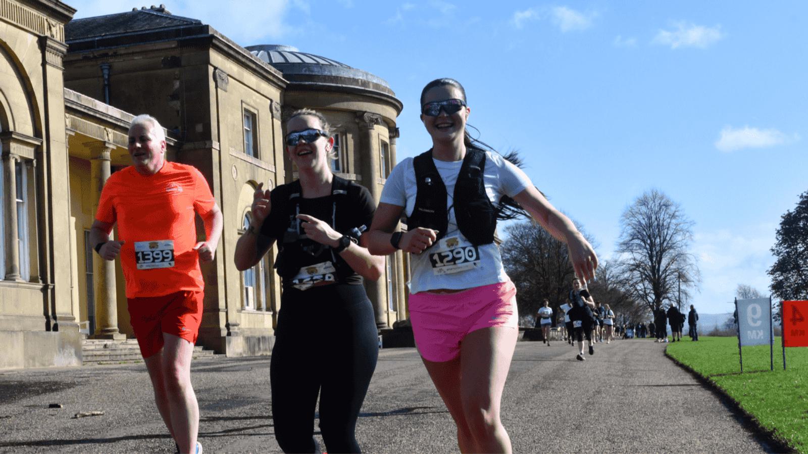 Three runners smile as they jog down a path alongside a historic building on a sunny day. The runner on the left wears an orange outfit, while the two on the right wear black, pink, and white. Other runners are visible in the background.