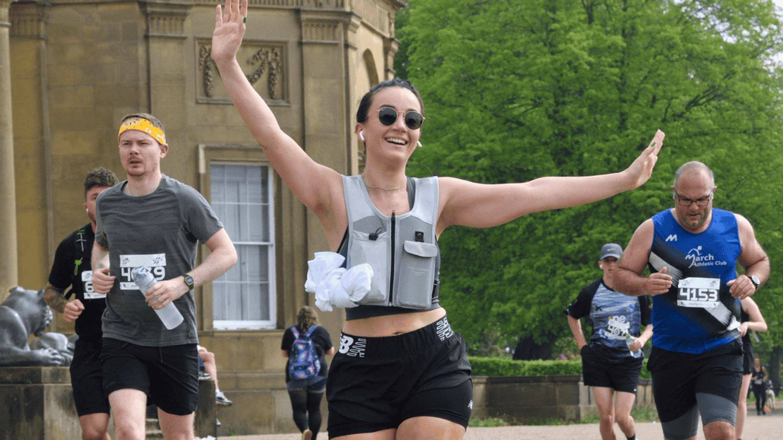 A woman in a weight vest smiles with arms raised, jogging alongside others in a running event. A man in a yellow headband runs nearby. They are in front of a historic building with lush green trees in the background.
