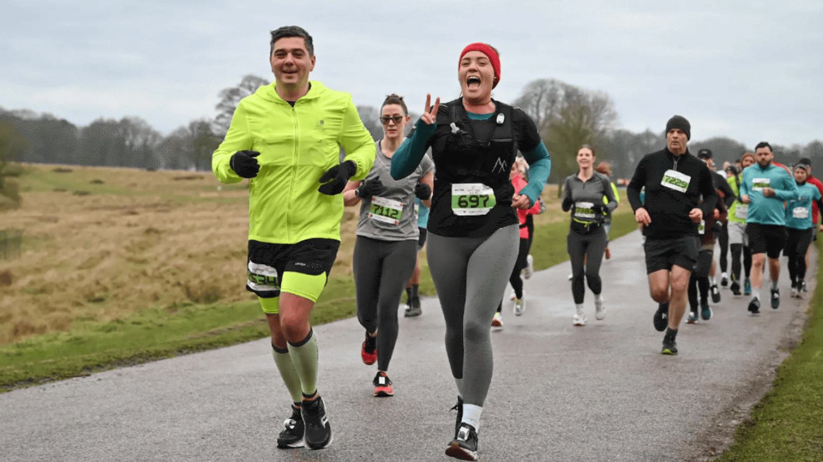 A group of runners in athletic gear participates in a marathon on a paved path. The lead female runner, wearing a red beanie and smiling, gestures a peace sign. Surrounding them is a park landscape with grass and trees under an overcast sky.