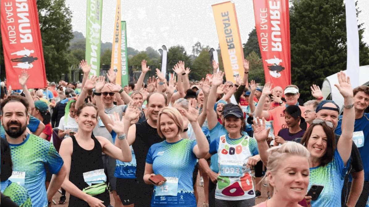 A large group of enthusiastic runners wave at the camera before a race. They are wearing colorful athletic gear and are surrounded by banners with text. Trees and more participants are visible in the background.