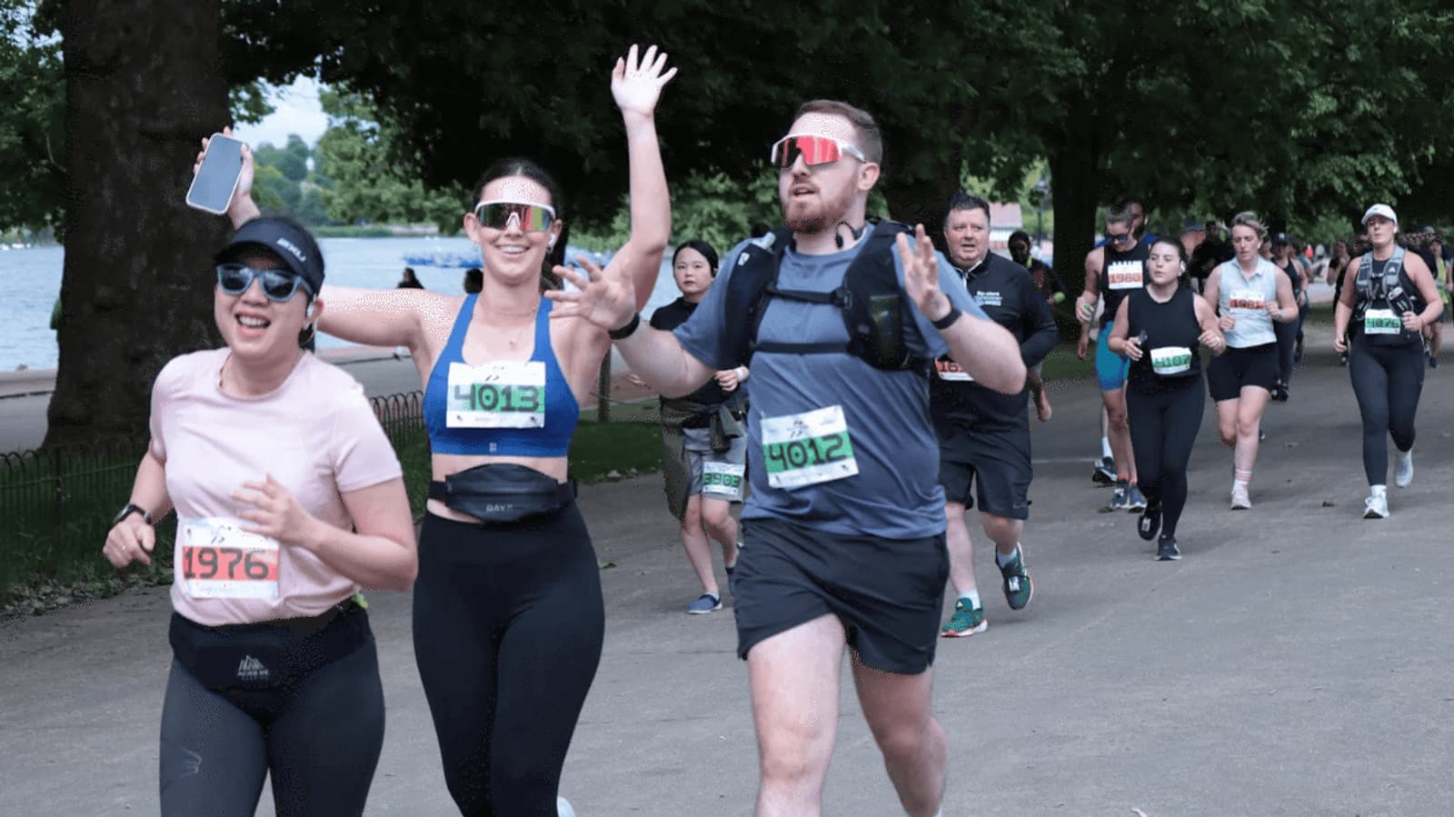 Participants in a race are running along a tree-lined path. Many are smiling and waving, while wearing numbered bibs and sunglasses. The atmosphere is energetic and cheerful, with a mix of men and women enjoying the event.