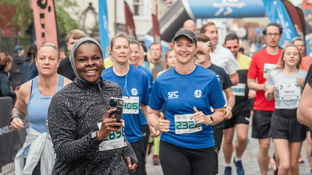 A group of people running in a race with numbered bibs. A woman in the front smiles broadly and holds a phone. Several participants wear blue shirts, and an inflatable arch is visible in the background.