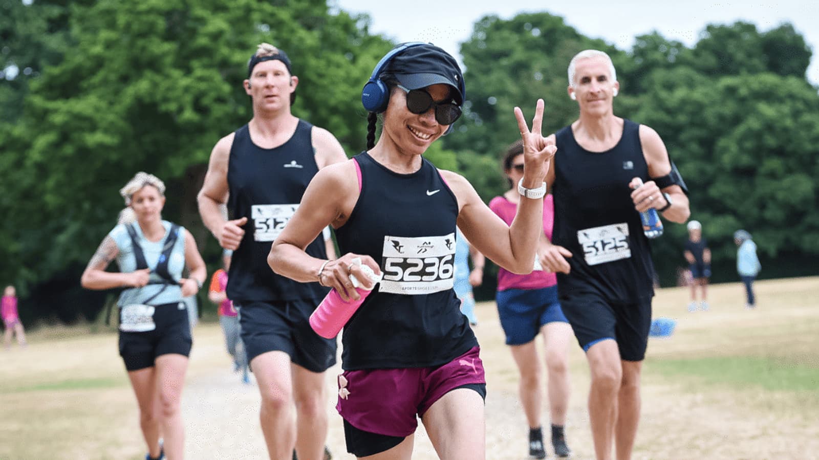 A group of runners in sportswear participating in a race. A person in the foreground is smiling, holding a pink water bottle, and making a peace sign. The background shows trees and a mix of other runners.