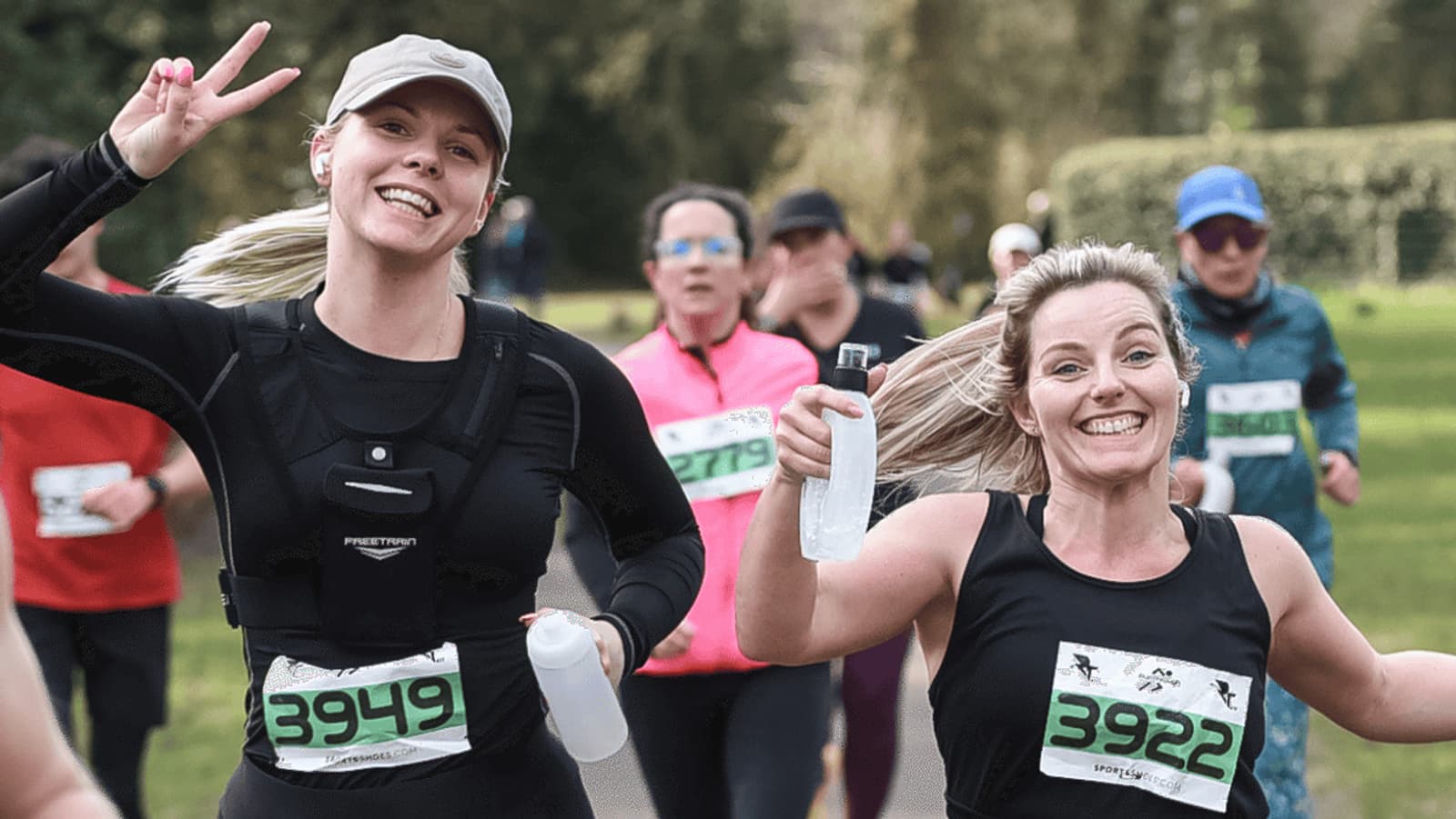 Two women in athletic wear, running a race with numbered bibs 3949 and 3922. They are smiling, one making a peace sign, both holding water bottles. Other runners are in the background on a tree-lined path.
