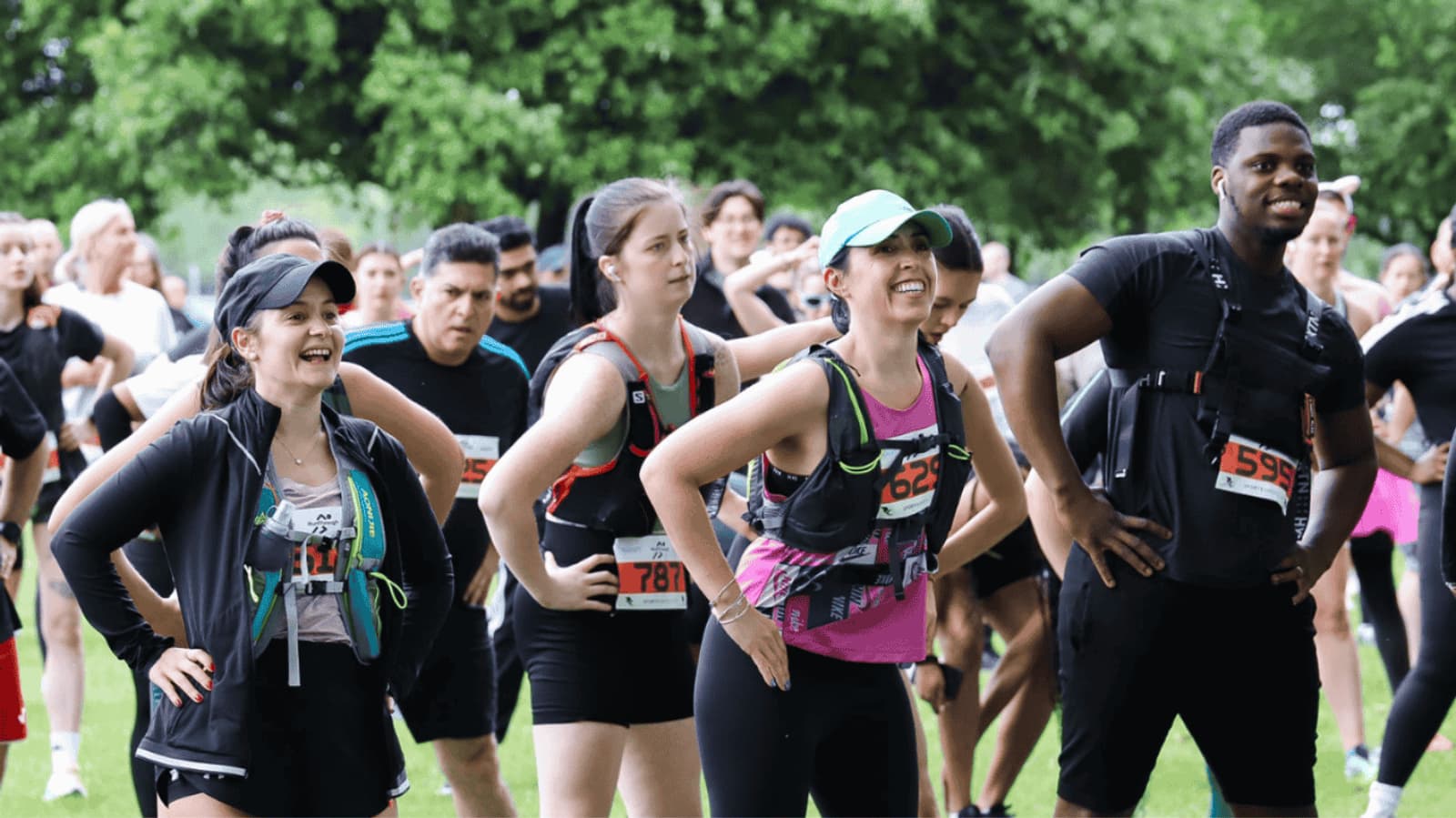 A diverse group of runners with race bibs gather outdoors, smiling and warming up in a park. They are dressed in athletic gear, and a green forested background is visible. The atmosphere is lively and energetic.