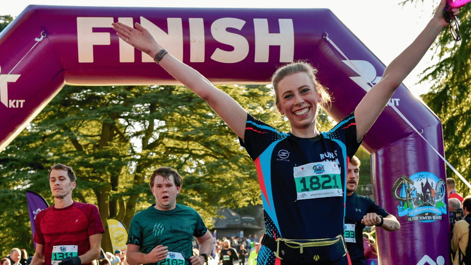 A woman with a race bib number 1821 joyfully crosses the finish line with her arms raised in victory. Behind her, other runners are approaching the finish. The scene is outdoors with trees and a large purple "FINISH" banner overhead.