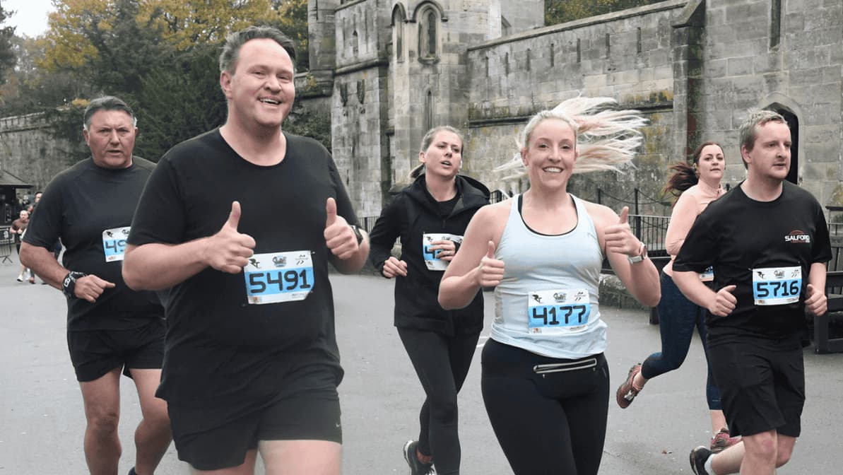 Runners participating in a race, wearing numbered bibs. A man and woman in the foreground are smiling and giving thumbs up. They are running alongside others on a street with a stone building in the background.