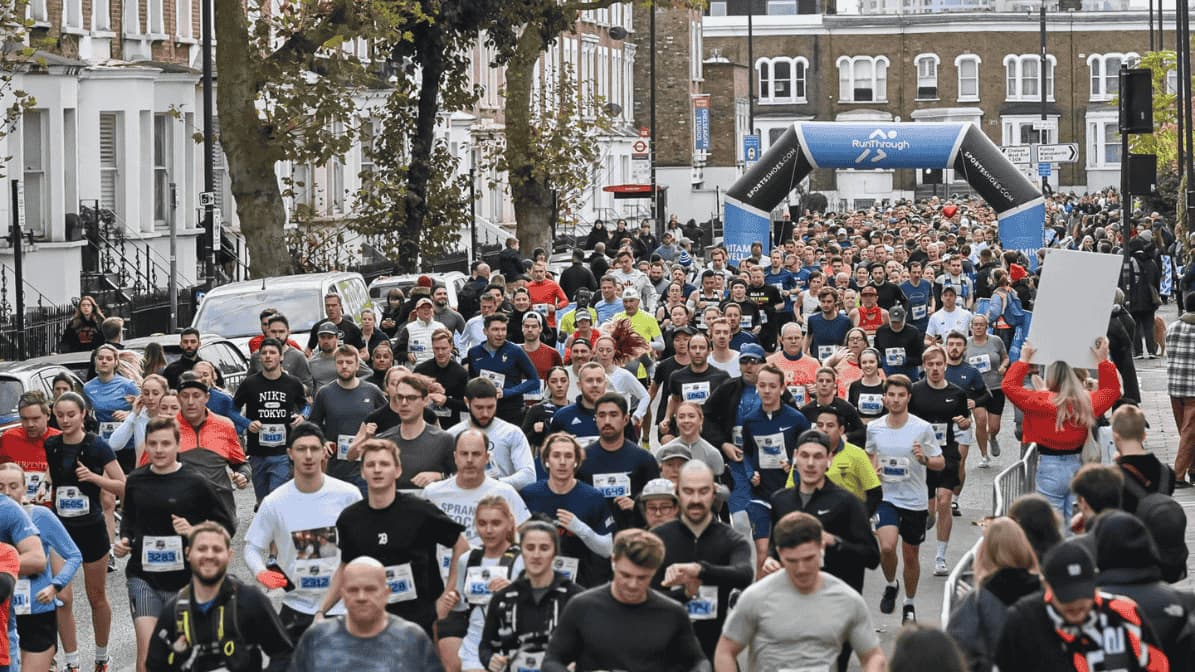 A large group of runners participating in a marathon on a city street. An archway with event branding stands in the background. Spectators line the sidewalk, and trees and buildings frame the scene.