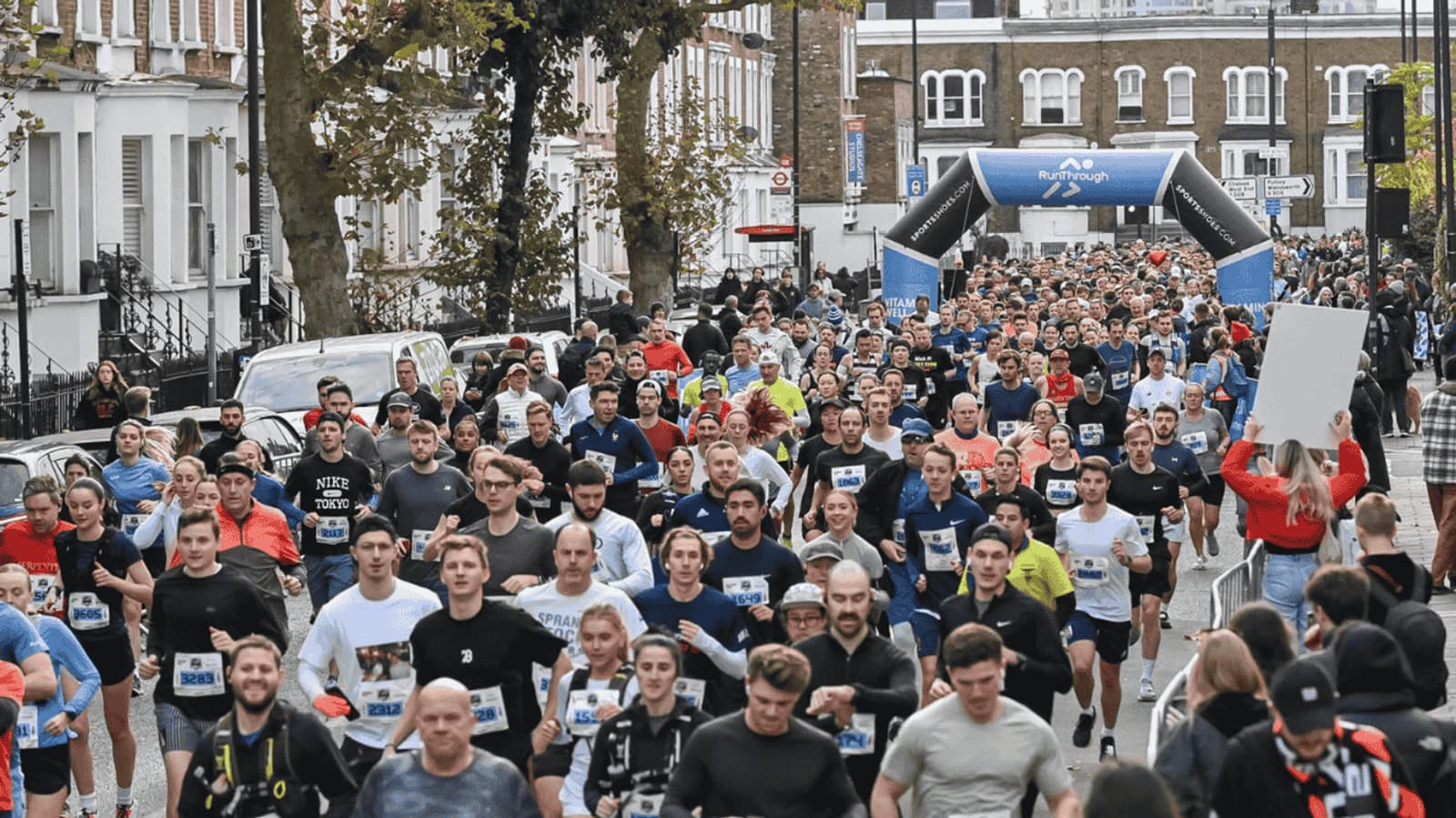 A large group of runners participating in a marathon on a city street. An archway with event branding stands in the background. Spectators line the sidewalk, and trees and buildings frame the scene.