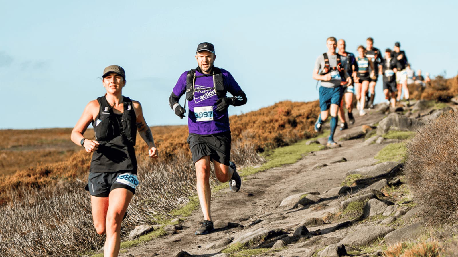 A group of people are running on a rocky trail through a grassy landscape. The sky is clear and blue. The runners, wearing athletic gear and numbered bibs, are moving in a line along the path. The terrain is rugged and open.