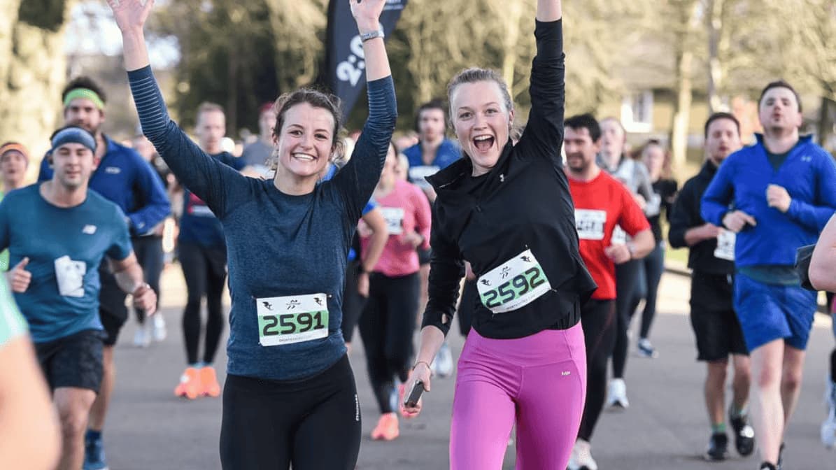 Two women in athletic gear, wearing numbered race bibs, smile and raise their arms triumphantly while running in a crowded outdoor race. Other participants are visible in the background. The event takes place on a sunny day.
