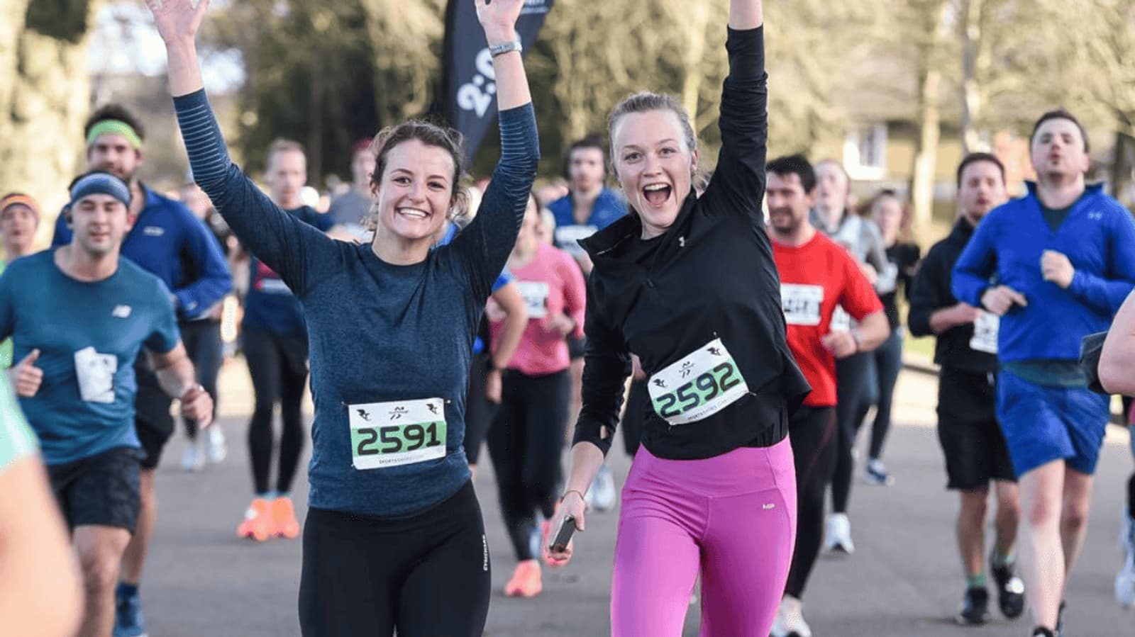 Two women in athletic gear, wearing numbered race bibs, smile and raise their arms triumphantly while running in a crowded outdoor race. Other participants are visible in the background. The event takes place on a sunny day.