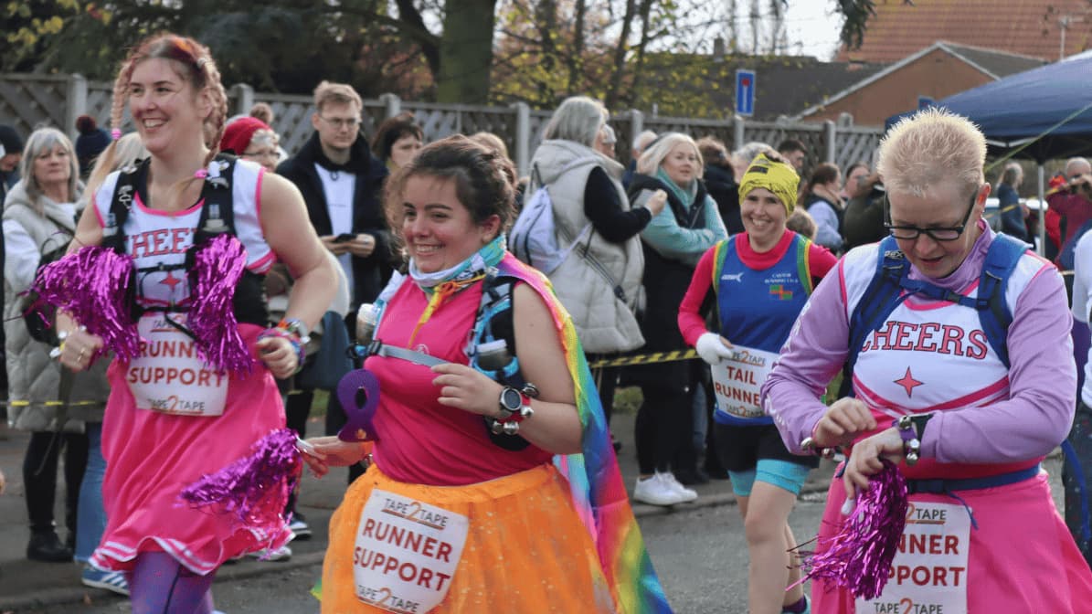 Three people with "Runner Support" signs cheer during a marathon. They're dressed in colorful outfits with pink tutus and pom-poms. A crowd watches in the background under a cloudy sky.