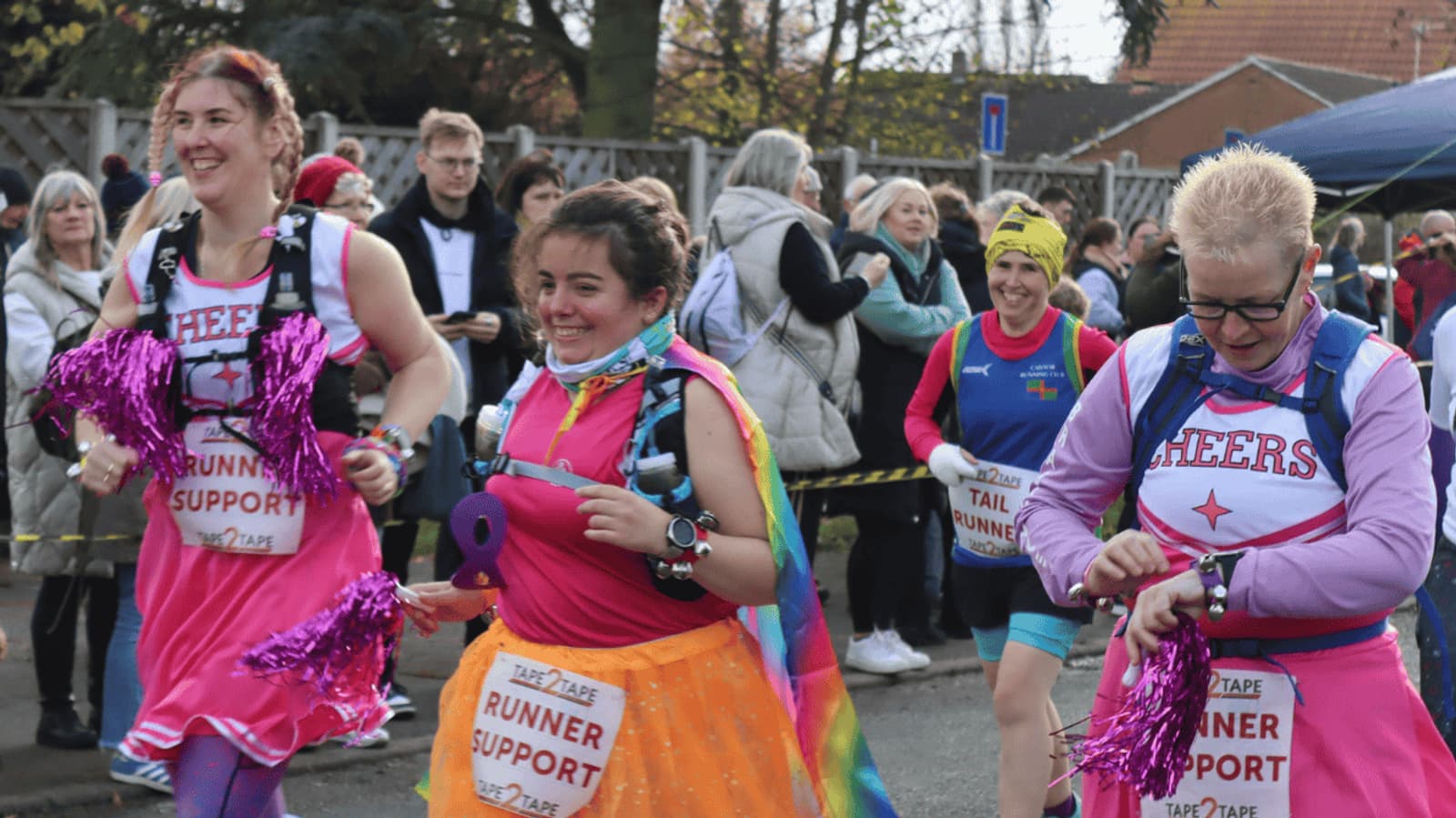 Three people with "Runner Support" signs cheer during a marathon. They're dressed in colorful outfits with pink tutus and pom-poms. A crowd watches in the background under a cloudy sky.