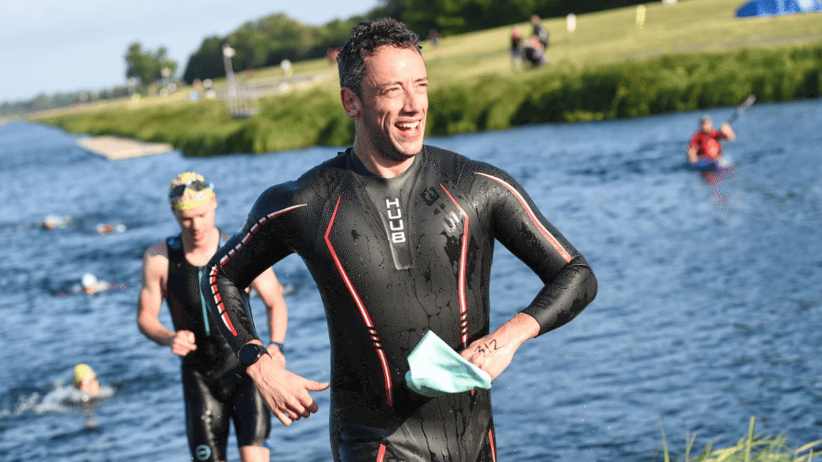 A person in a wetsuit is emerging from a lake, smiling and holding a swim cap. Other swimmers in the background are still in the water. The scene is sunny with green grass and trees visible.