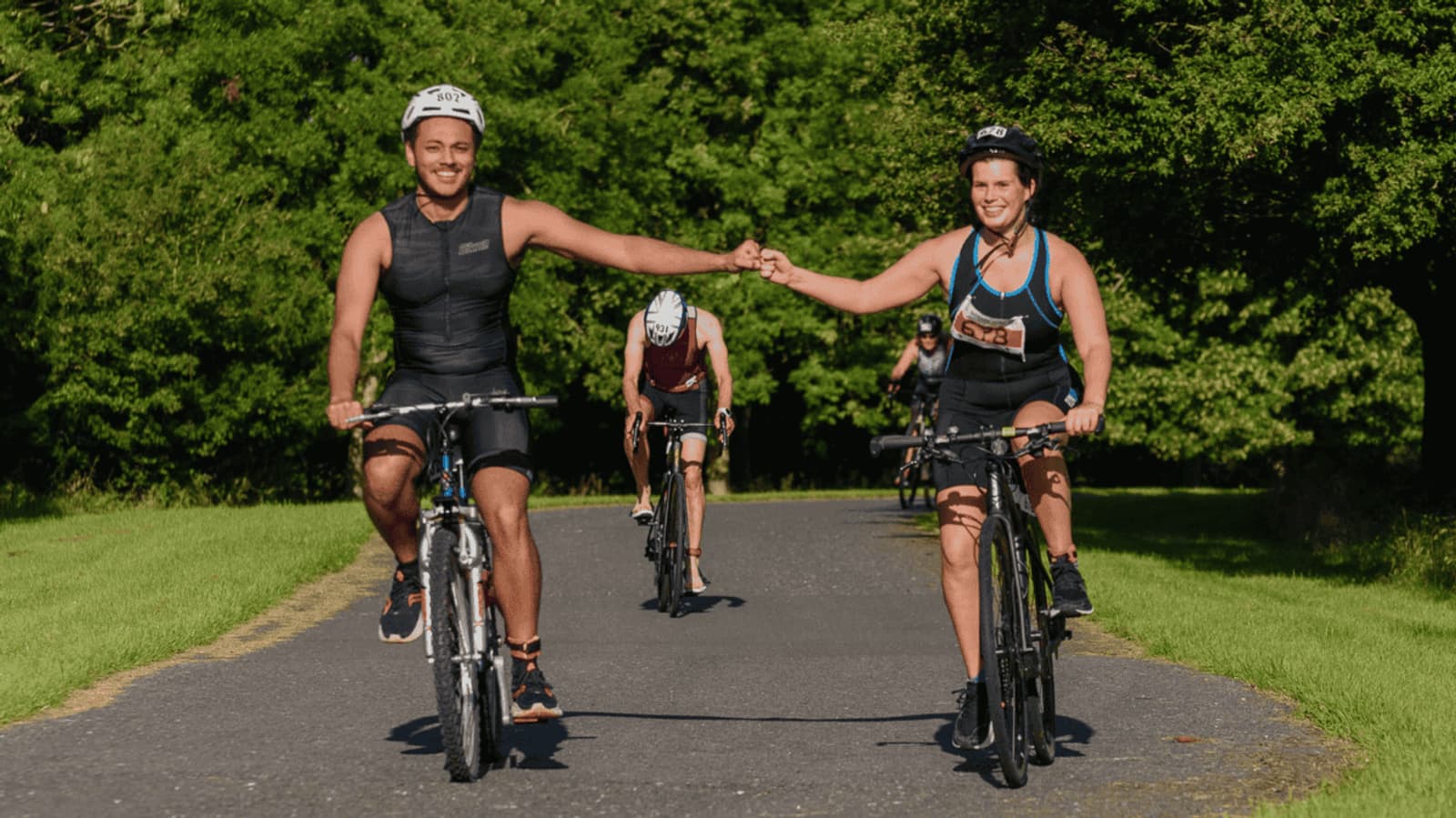 Two cyclists fist-bumping while riding on a paved path in a park, wearing helmets and athletic gear. They are followed by two more cyclists in the background. Lush green trees and grass line the path on a sunny day.