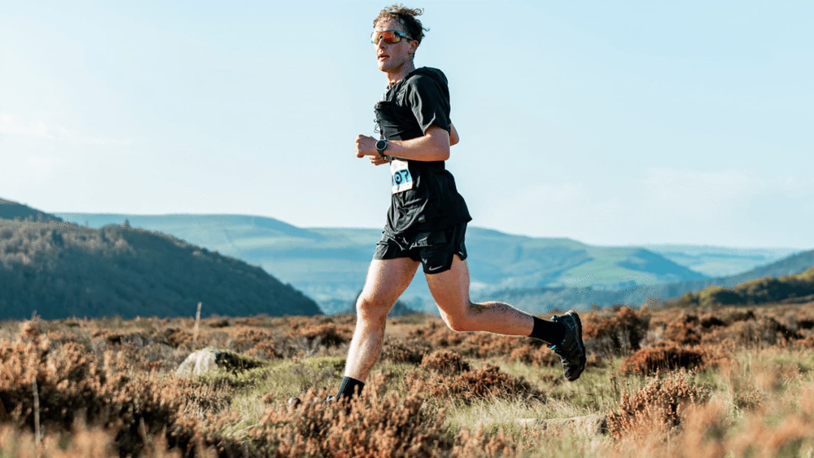 A person is running in a scenic, open landscape. They are wearing sunglasses, a black shirt, and shorts, with a race number on their waist. The background features hills, a blue sky, and scattered vegetation.