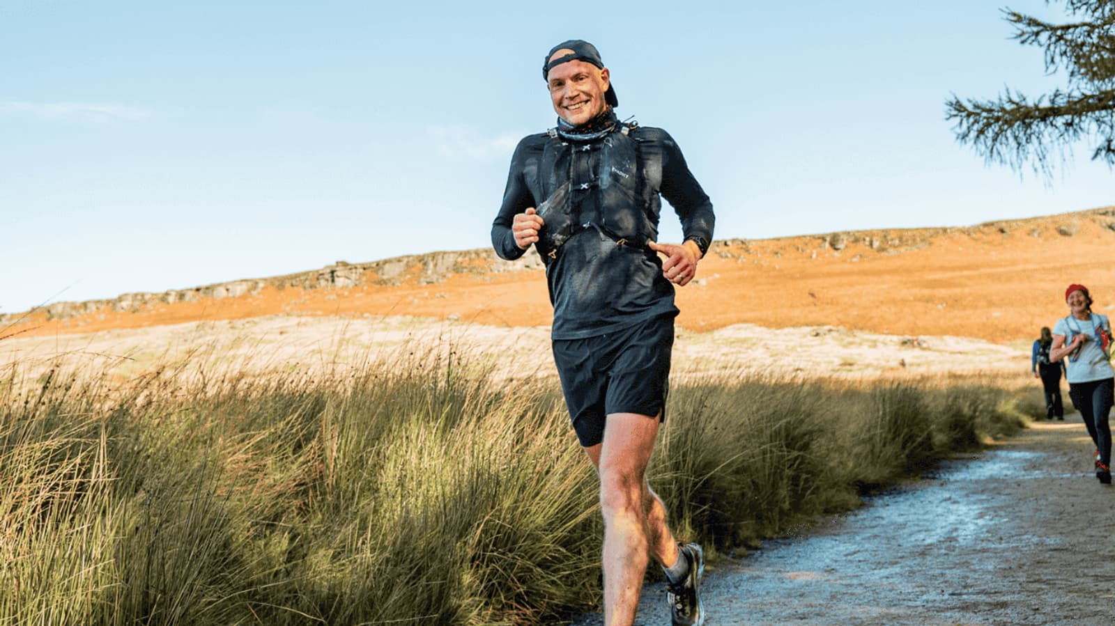 A person in a black running outfit and cap jogging along a trail in a grassy landscape under a clear blue sky. Other runners are visible in the background. The sun casts a warm light on the scene.