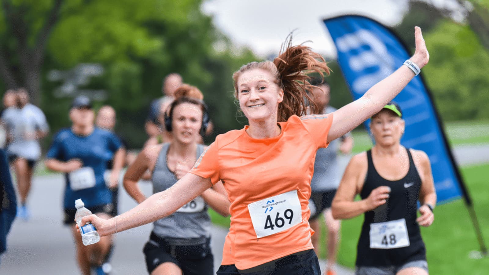 A woman in an orange shirt, with number 469, smiles and raises her arms while running in a race. Other runners are visible in the background, including a woman wearing number 48. The scene is outdoors with greenery.
