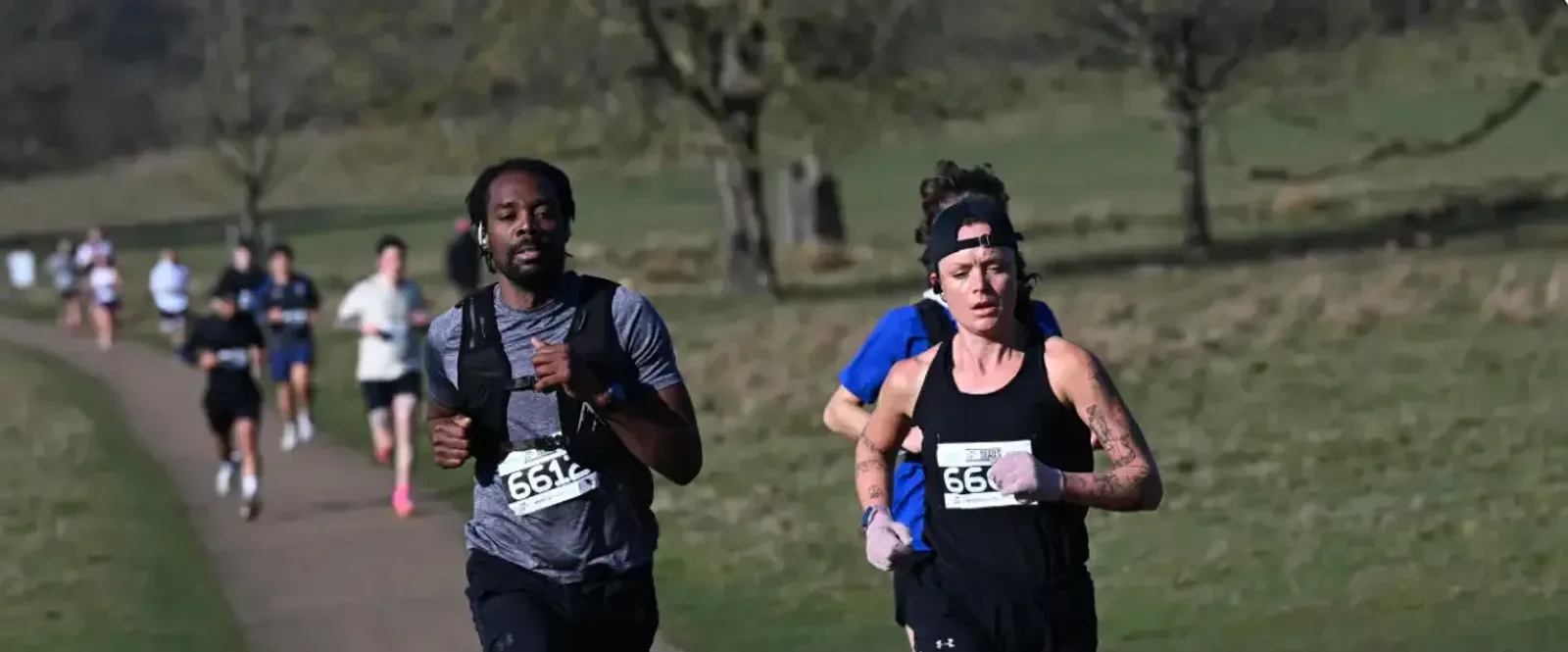 Two runners, a man and a woman, jog on a park path during a race, wearing numbered bibs. More competitors run behind them on a sunny day with grass and trees in the background.