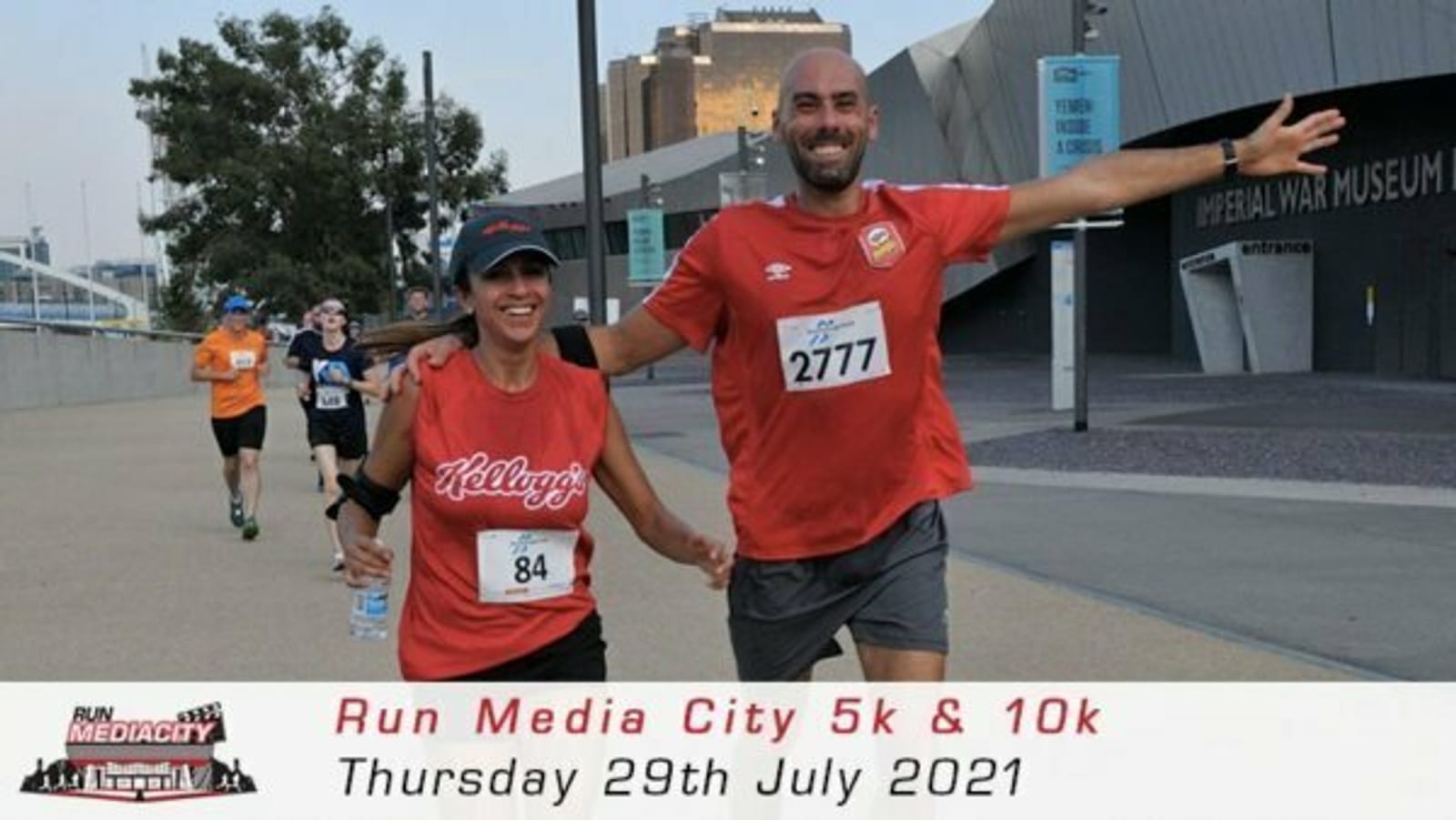 Two runners, a man and a woman, joyfully hold hands while running near the Imperial War Museum. Both are wearing red tops; the man (bib number 2777) is bald, and the woman (bib number 84) has a cap. Other runners are visible in the background. Text on the image reads "Run Media City 5k & 10k, Thursday 29th July 2021.