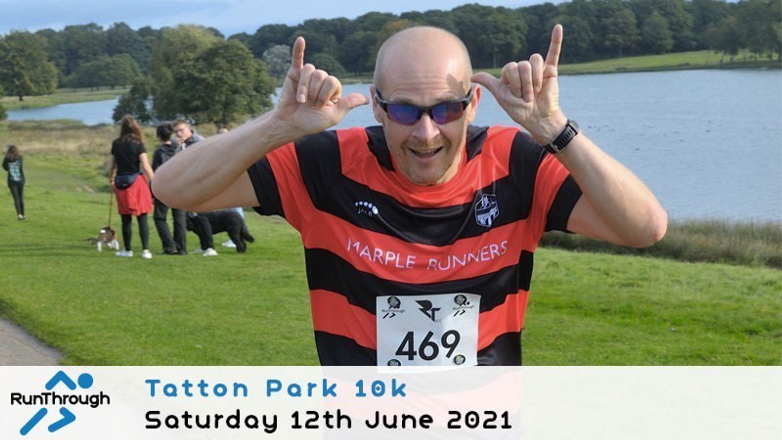 A smiling runner in a black and red striped shirt makes a playful gesture with both hands during the Tatton Park 10k race, held on Saturday, 12th June 2021. Other people and a lake are visible in the background.