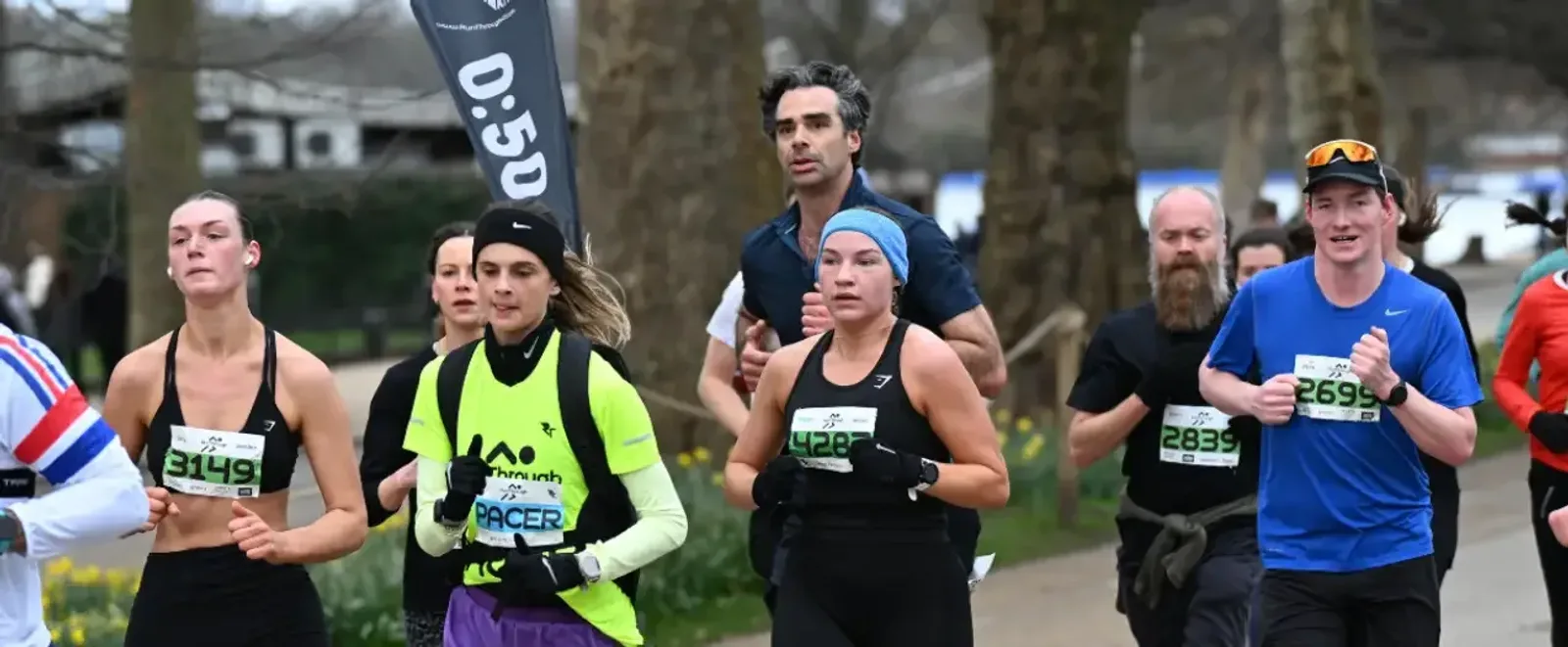 A group of runners participate in a race outdoors, wearing numbered bibs and athletic clothing, with trees and a "0:50" pacer flag visible in the background.