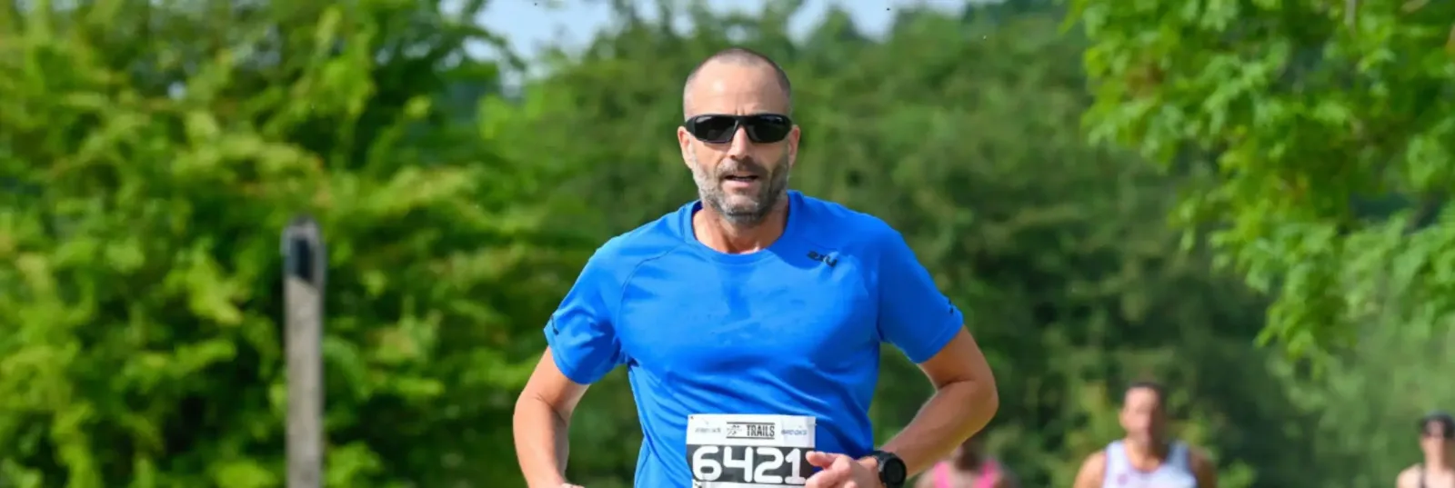 A man wearing sunglasses and a blue shirt runs outdoors during a race, with a race bib number 642 displayed on his shirt. Green trees and other runners are visible in the blurred background.