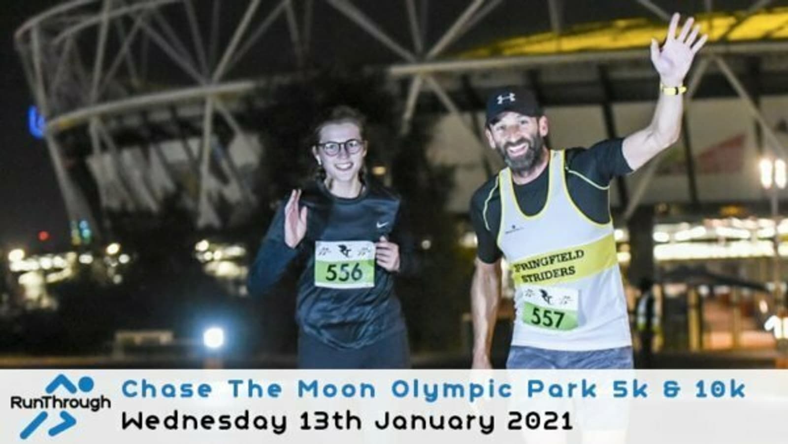 Two runners smile and wave at the camera during the Chase The Moon Olympic Park 5k & 10k event on Wednesday, 13th January 2021. They wear race bibs numbered 556 and 557. The background features a stadium with bright lights at night.