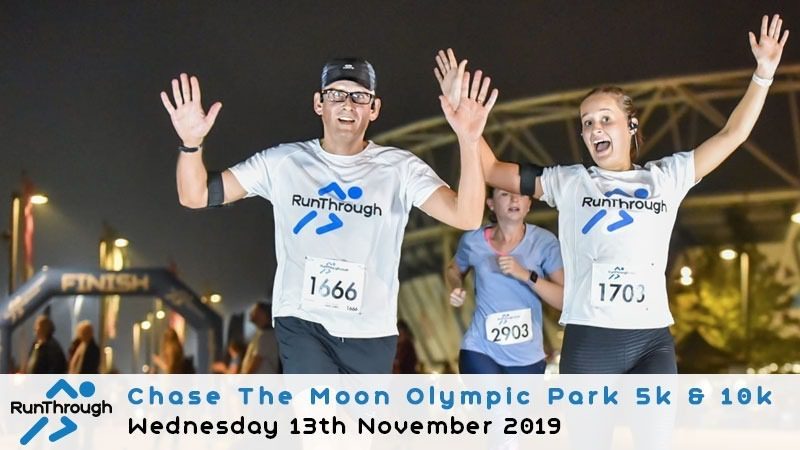 A group of runners nearing the finish line during the "Chase The Moon Olympic Park 5k & 10k" event on Wednesday, November 13th, 2019. Two runners in the foreground wear white RunThrough shirts with bib numbers 1666 and 1703, with the Olympic Stadium visible in the background.