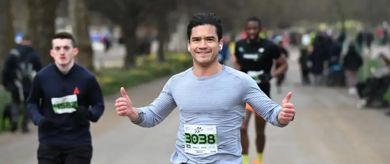A man in a gray long-sleeve shirt smiles and gives two thumbs up while running a race in a park, with other runners and trees visible in the background.