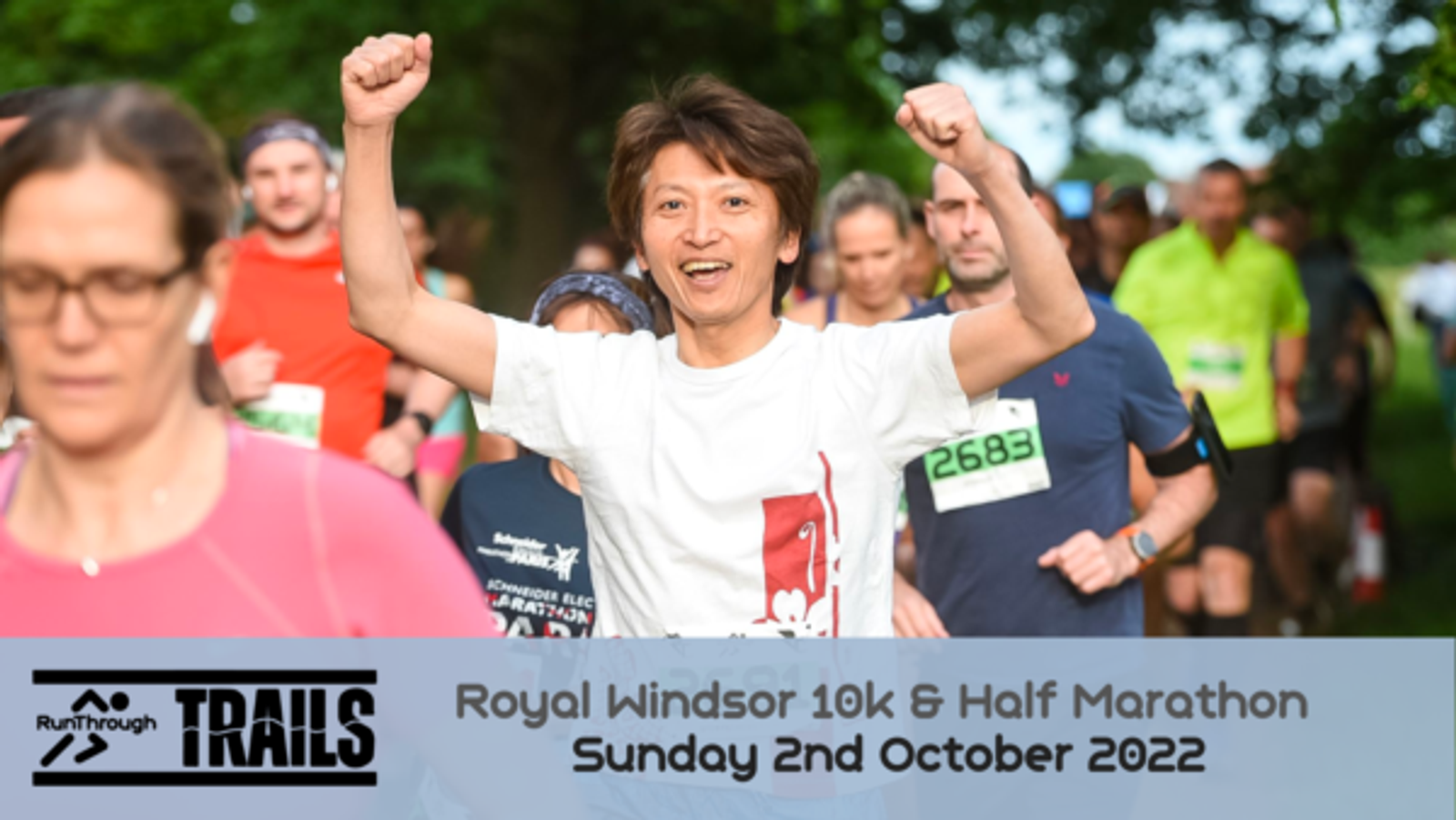 A group of runners participating in the Royal Windsor 10k & Half Marathon on Sunday, October 2nd, 2022. A person in the center, wearing a white shirt and smiling, has their arms raised in triumph. Other runners in colorful attire surround them.