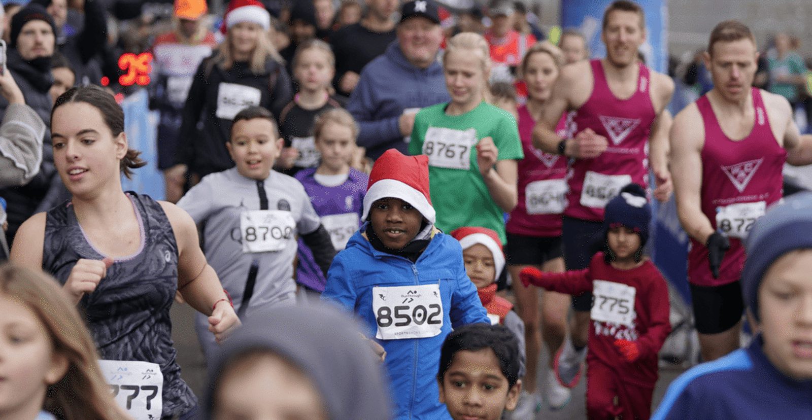 A group of runners, including adults and children, participate in a race. One child in front wears a blue jacket and a Santa hat. Many runners wear numbered bibs and athletic clothes. The atmosphere is energetic and festive.