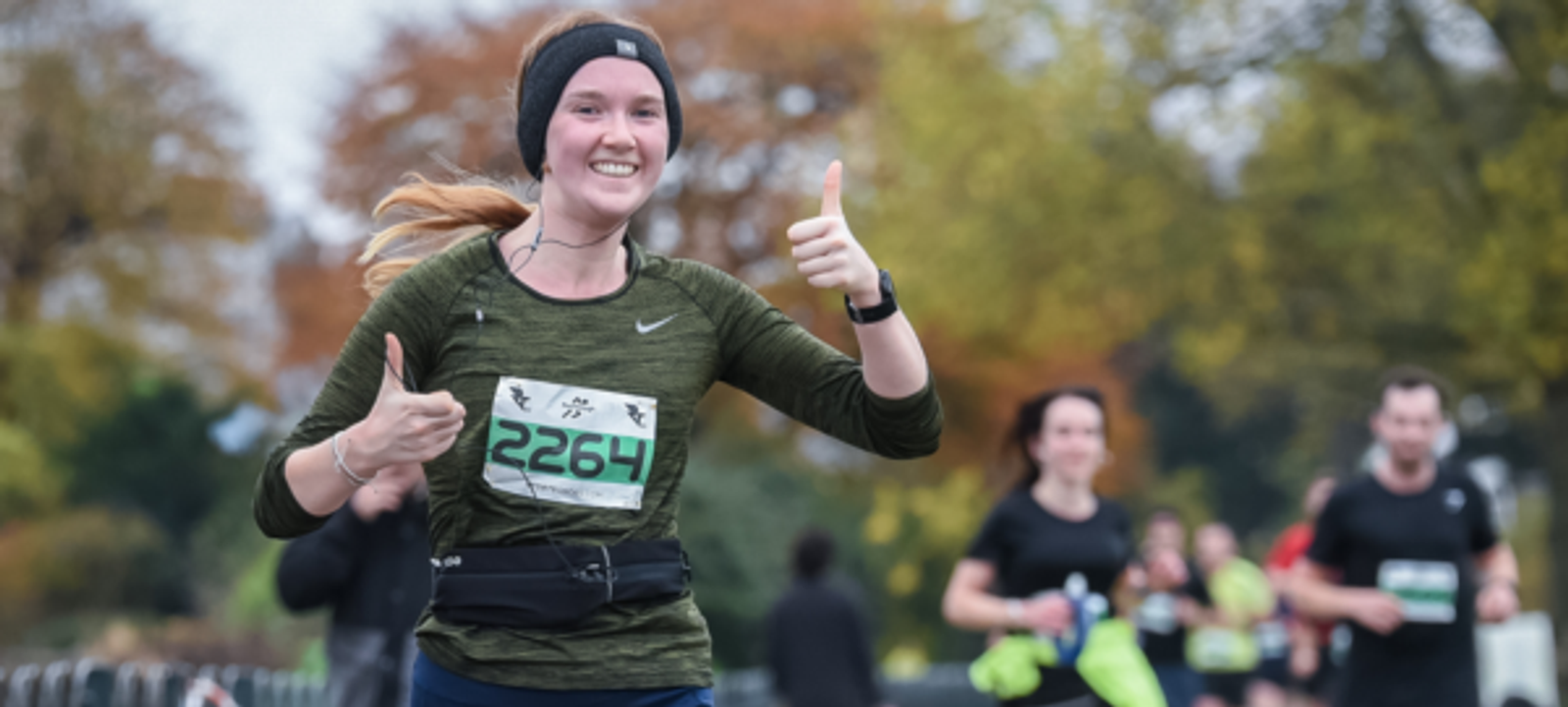 A runner wearing a green shirt, black cap, and race bib number 2264 gives a thumbs-up during a race. Blurred participants and autumn trees are in the background. Text overlay reads "Greenwich 5k, 10k & Juniors, Saturday 5th October 2024" with the RunThrough logo.