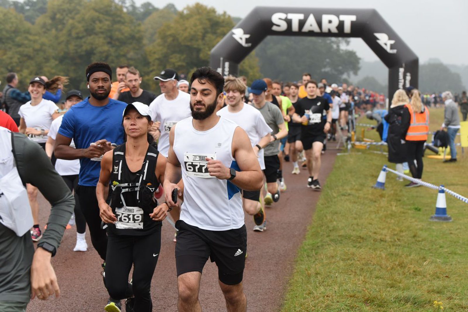 Runners participate in a race, starting from an arch that reads "START." Participants wear race numbers and various athletic outfits. The background features trees and a foggy sky. The path is lined with blue cones and tape, and a few spectators and volunteers are visible.