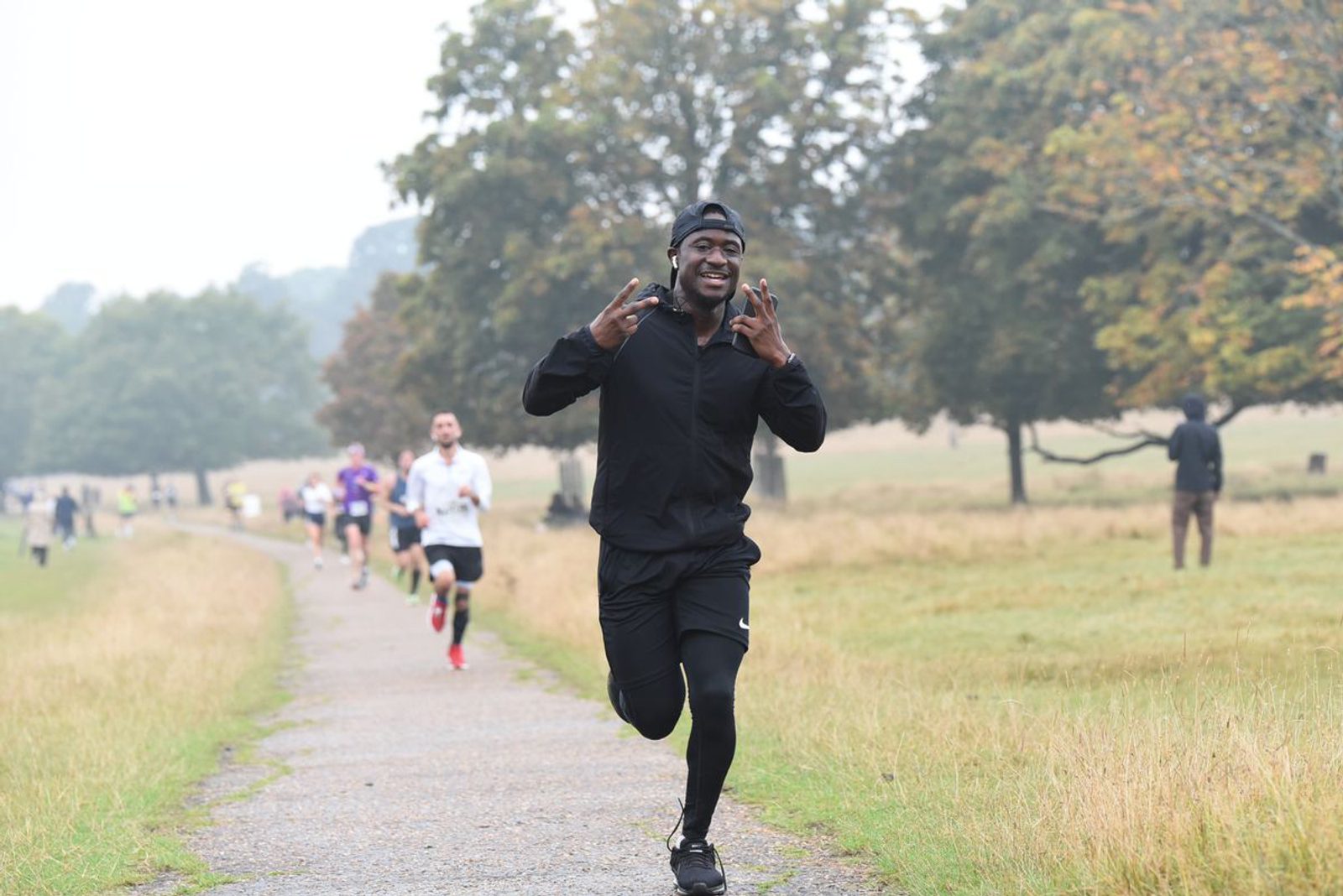 A person wearing a black outfit and cap is running on a pathway in a park, smiling broadly and making a peace sign with both hands. Other runners are visible in the background. The park has grass on either side of the path and trees with lush green foliage.