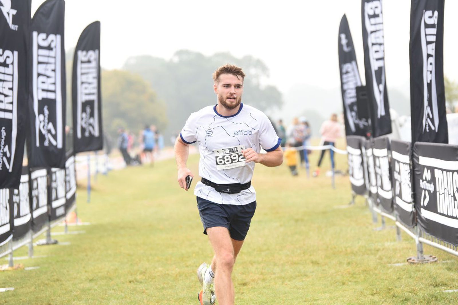 A man wearing a white shirt and black shorts is running in a race, holding a wristband with the number 6895. He is on a grassy path lined with black flags that read "TRAILS." Other runners and spectators are visible in the background.