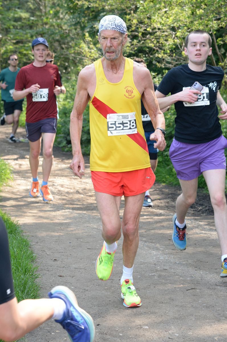 A group of people running in a race on a trail through a wooded area. Prominent in the frame is an older man wearing a yellow and red tank top with the number 5538, and bright yellow shoes. He is surrounded by other runners in various athletic attire.