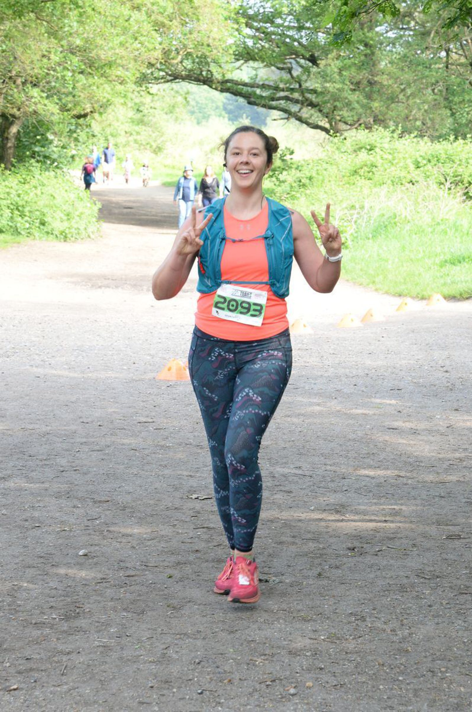 A woman wearing a coral tank top, patterned leggings, and pink running shoes smiles and displays peace signs with both hands while running on a tree-lined path during a race. She wears a race bib with the number 2093 pinned to her shirt, and other runners are visible in the background.