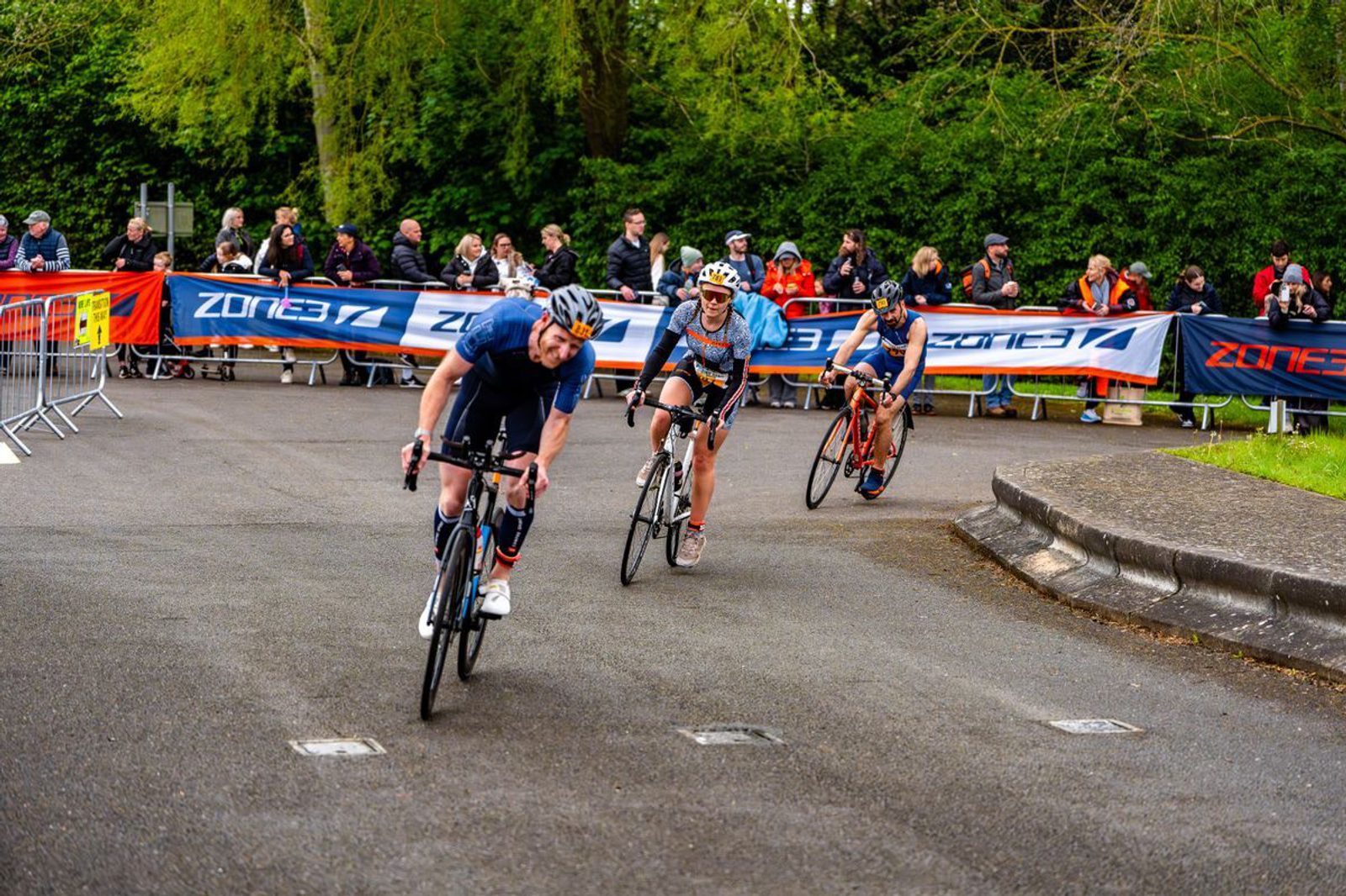Three cyclists race on a paved road with spectators behind barriers watching them. Two cyclists are closely competing beside each other in the lead, while the third follows slightly behind. The scene is set in a lush, green outdoor area.