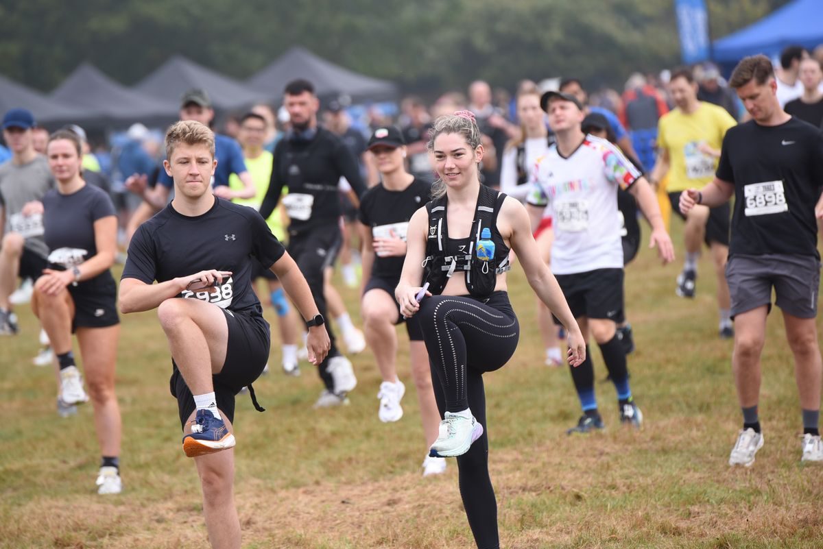 A group of people participates in a pre-race warm-up routine outdoors. They are lifting one knee high while balancing on the other leg. They are dressed in athletic gear, and all appear focused and engaged. The event tents and more participants are visible in the background.