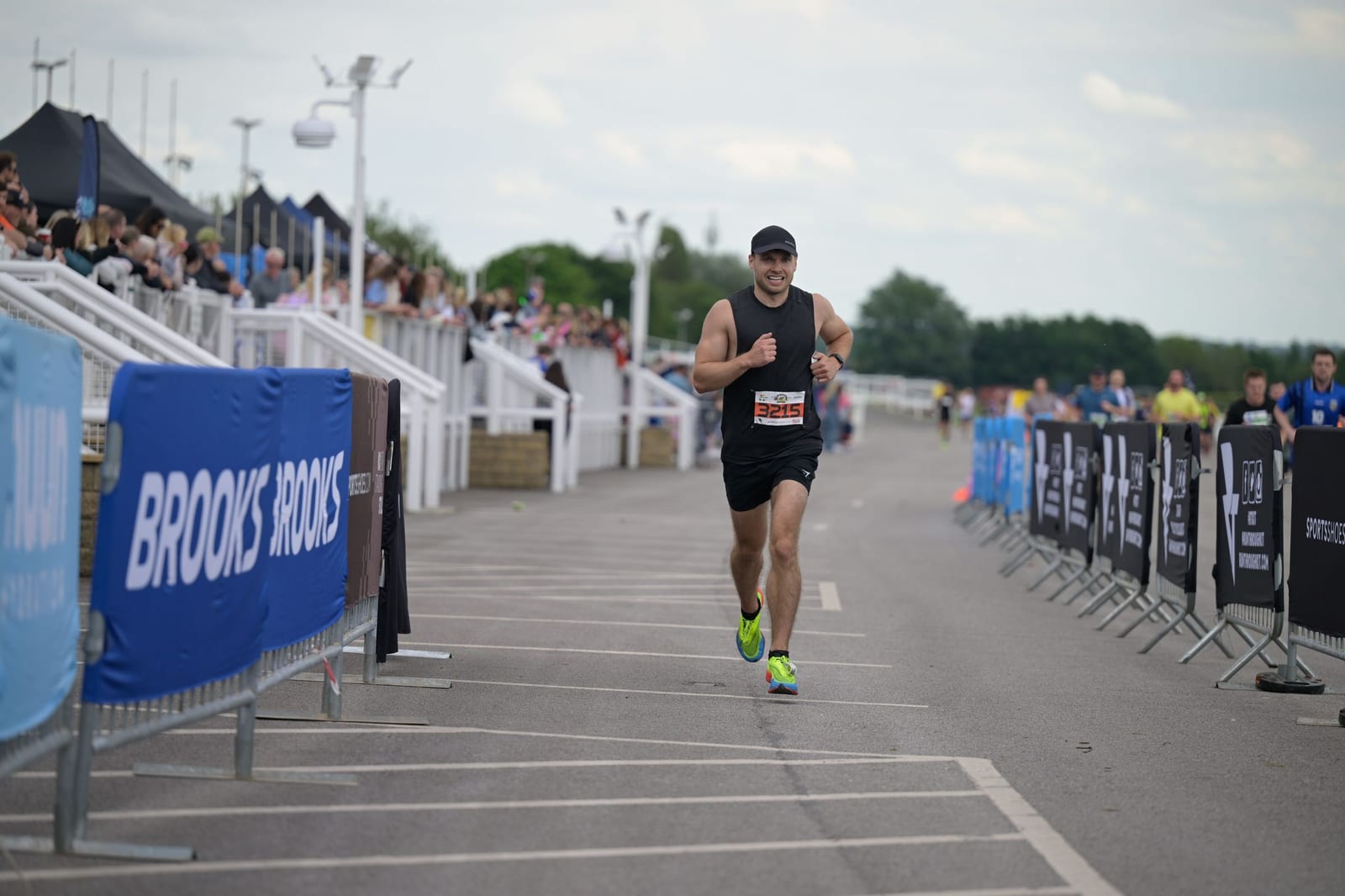 A runner wearing a black tank top, black shorts, and bright yellow-green running shoes is participating in a race. The runner is on a road with barriers and sponsorship banners on both sides, with spectators watching from the stands in the background.