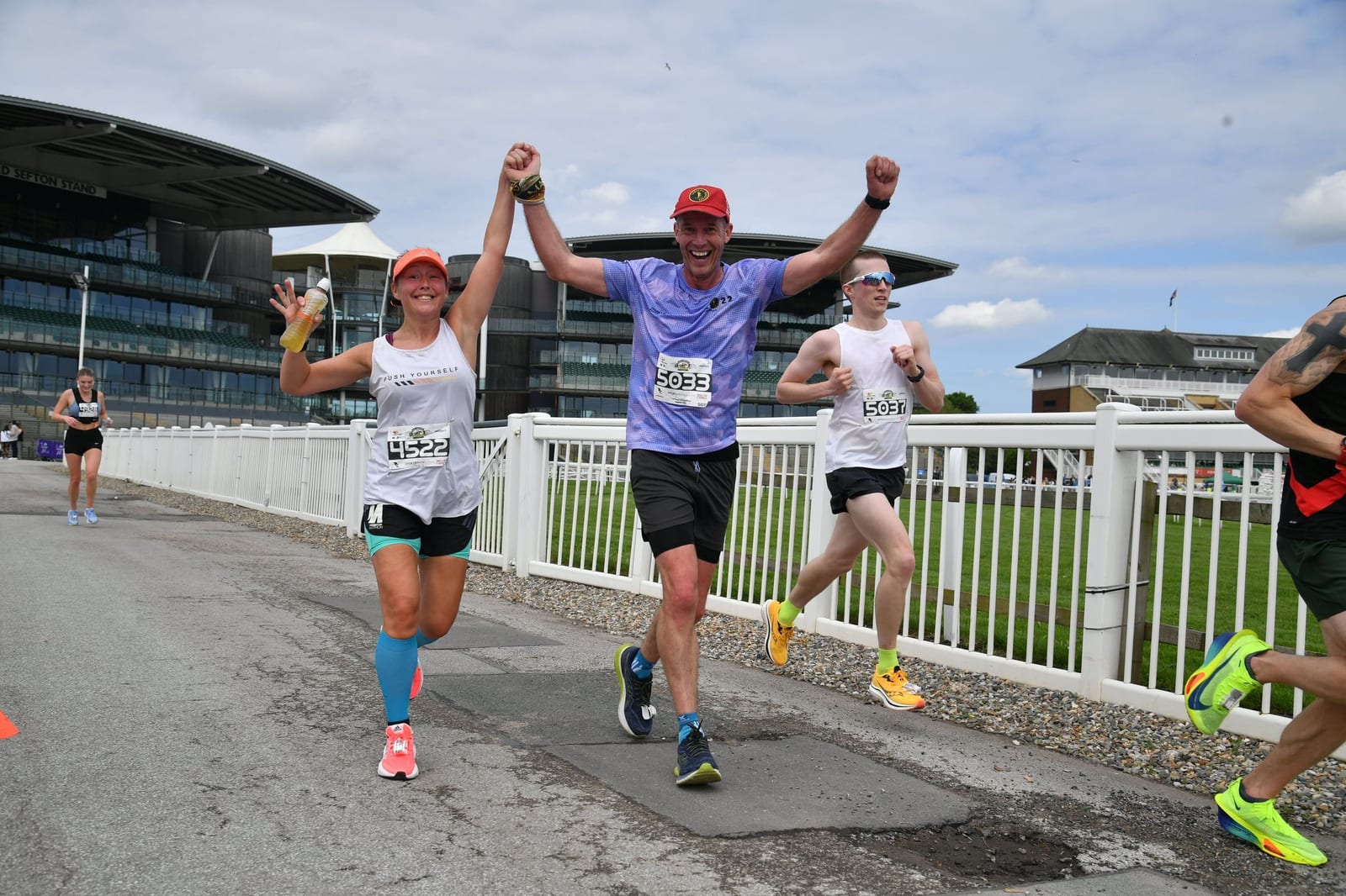 Three runners are participating in a race, with two of the runners in the foreground holding hands and smiling. One is wearing a blue headband and orange shoes, while the other is in a purple shirt and black shorts. The race track is near a modern, large building.